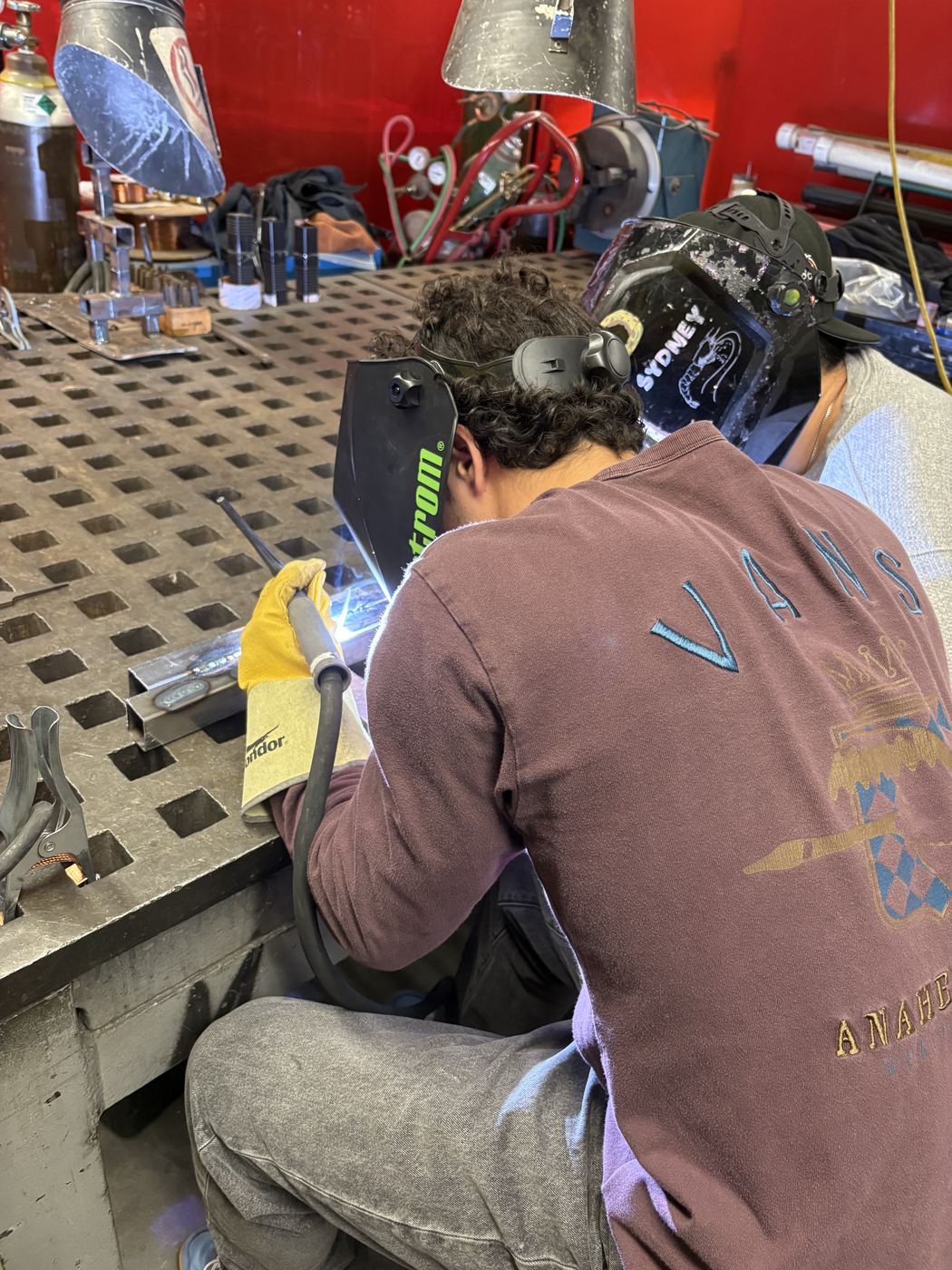 TIG welding at the fabrication table with a second team member alongside.